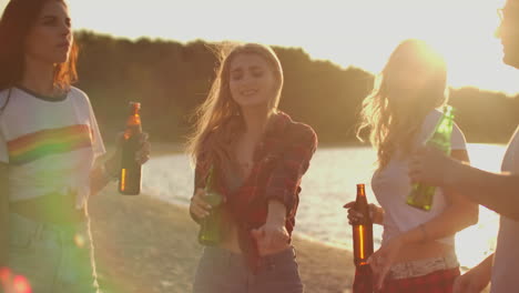 Three-hot-girls-are-dancing-in-short-t-shirts-and-shorts-with-beer-on-the-sand-beach-at-sunset.-Their-hair-is-flying-on-the-wind.